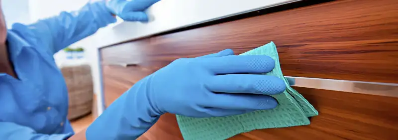 Cleaner wearing blue gloves wiping a wooden cabinet drawer with a microfiber cloth for dust and grime removal, Ogden, UT.