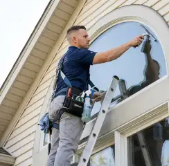 Professional window cleaner on a ladder using a squeegee to clean an exterior arched window, Ogden, UT.